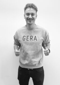 Junger Mann trägt einen Pullover mit der Aufschrift "Gera", lacht und zeigt beide Daumen nach oben
