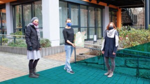 Drei Frauen stehen mit Abstand und Maske nebeneinander, die mittlere Frau hält ein verpacktes Weihnachtsgeschenk in die Kamera