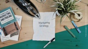 Schreibtisch mit Büchern, Stiften, einer Pflanze, einem Zettel, auf dem "Marketing Strategy" steht und einem Bügeleisen, das den Zettel glatt bügelt
