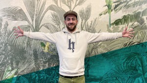Mann mit beigem Oberteil und Cap steht mit ausgebreiteten Armen vor einer Tapete im Dschungel-Design