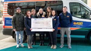 Fünf Frauen und zwei Männer stehen vor einem Kleinbus und halten einen großen Spendenscheck in die Kamera