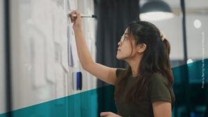 Junge Frau mit dunklen Haaren und Brille schreibt etwas an ein Whiteboard, an dem viele Zettel hängen