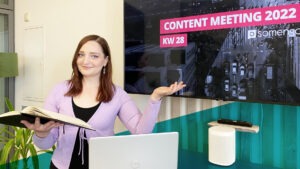 Junge Frau mit braunen Haaren posiert vor einem Fernseher und hält ein Notizbuch in einer Hand