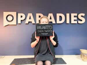 Blonde Frau sitzt vor einer Wand mit dem Schriftzug "Paradies" und hält ein Schild mit Aufschrift vor ihr Gesicht
