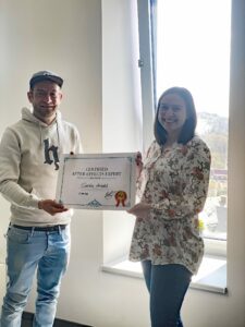 Ein Mann und eine Frau stehen nebeneinander, halten in der Mitte ein großes Papier mit einem Zertifikat und lächeln in die Kamera