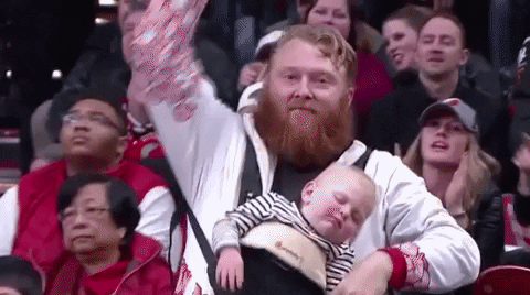 Mann mit Vollbart tanzt und zeigt enthusiastisch auf sein schlafendes Baby, das er in einem Tragegurt trägt