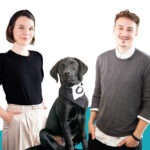 Eine weibliche und eine männliche Person sowie ein schwarzer Hund posieren vor einem blau-weißen Hintergrund