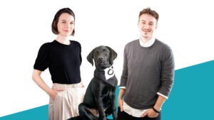 Eine weibliche und eine männliche Person sowie ein schwarzer Hund posieren vor einem blau-weißen Hintergrund