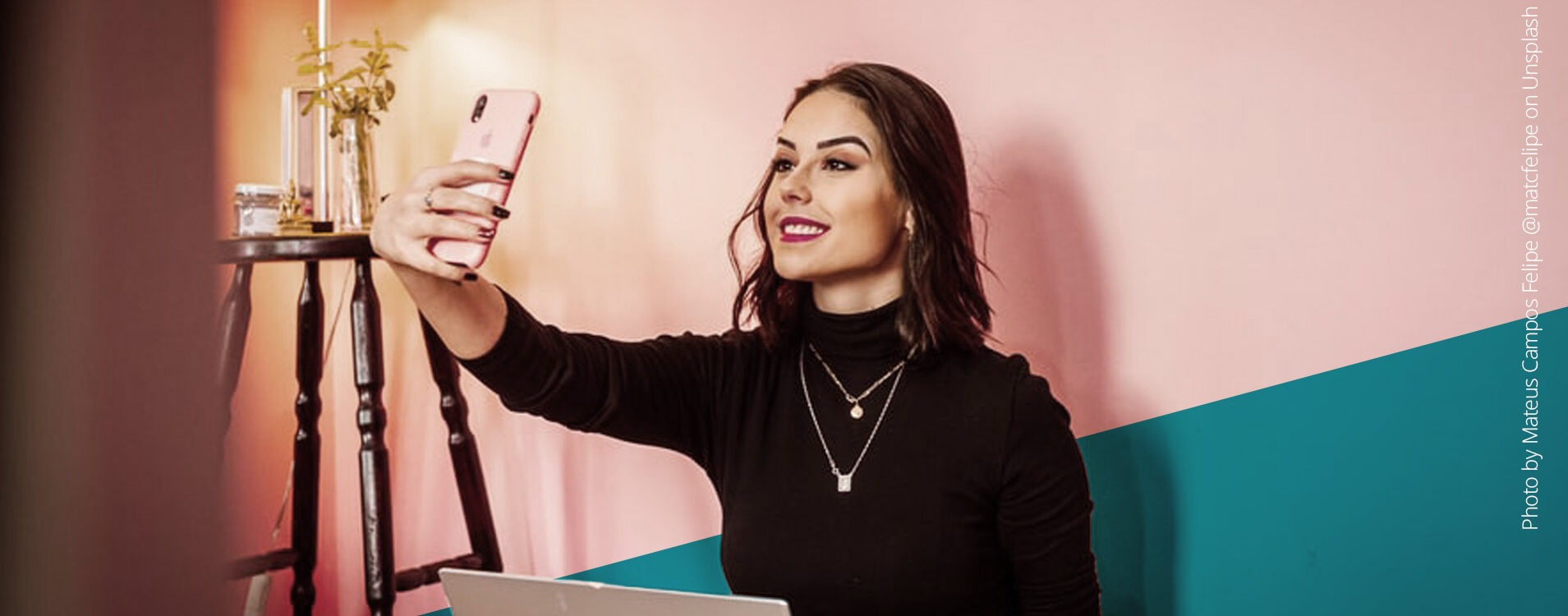 Junge Frau mit dunklen Haaren und Laptop auf dem Schoß macht ein Selfie vor einer pinken Wand