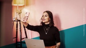 Junge Frau mit dunklen Haaren und Laptop auf dem Schoß macht ein Selfie vor einer pinken Wand