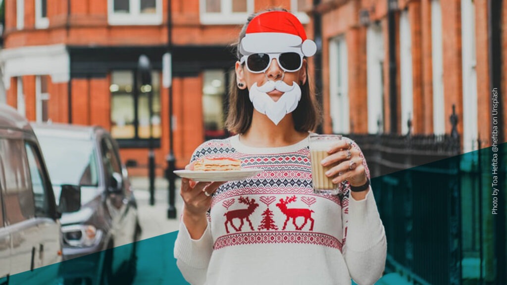 Frau mit Sonnenbrille, Weihnachtsmannmütze und weißem Kostüm-Bart, die einen Milchkaffe und ein Stück Kuchen in der Hand hält, steht auf der Straße