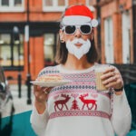 Frau mit Sonnenbrille, Weihnachtsmannmütze und weißem Kostüm-Bart, die einen Milchkaffe und ein Stück Kuchen in der Hand hält, steht auf der Straße