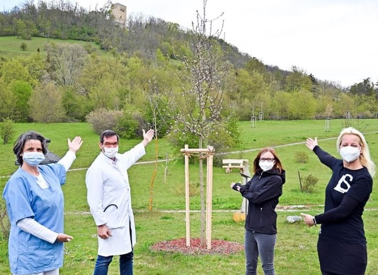 Vier Personen stehen draußen und zeigen auf einen frisch gepflanzten Baum, während sie in die Kamera schauen