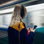 Frau mit blonden Haaren und Kopfhörern an einer U-Bahn-Station, durch die gerade eine Bahn fährt