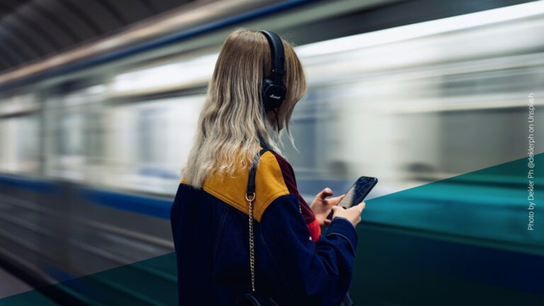Frau mit blonden Haaren und Kopfhörern an einer U-Bahn-Station, durch die gerade eine Bahn fährt