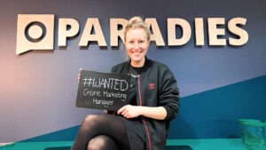 Junge blonde Frau sitzt vor einer Wand mit dem Schriftzug "Paradies" und hält eine Tafel, auf der steht "#wanted Online Marketing Manager"