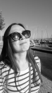 Junge Frau mit braunen Haaren und Sonnenbrille an einem Hafen schaut lächelnd nach oben in die Sonne