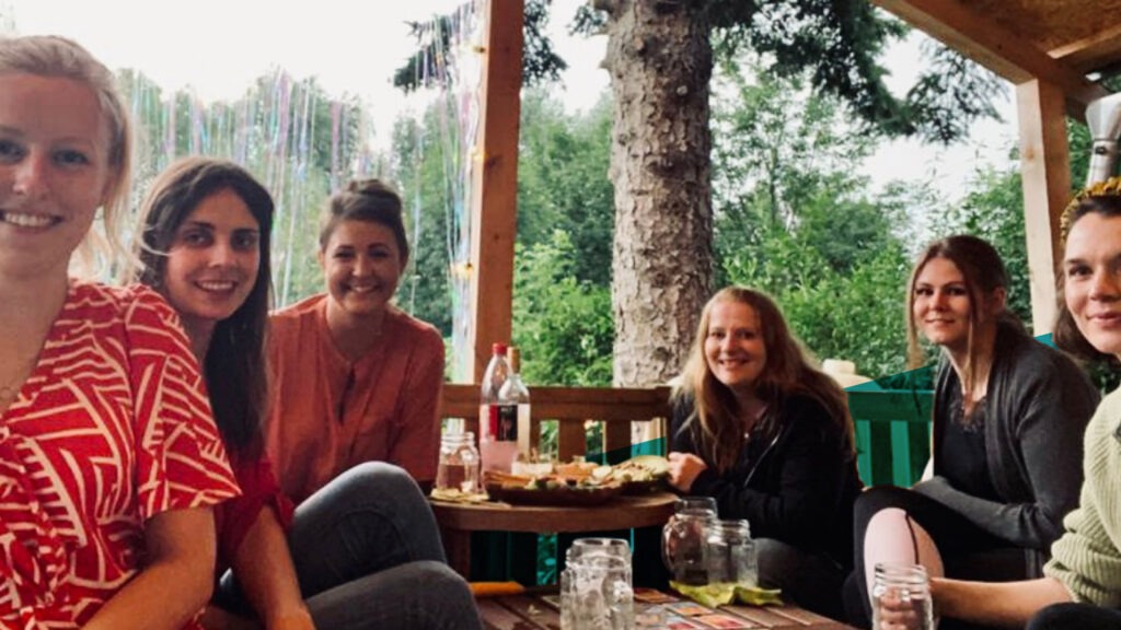 Sechs Frauen sitzen um einen Tisch im Garten und lächeln in die Kamera