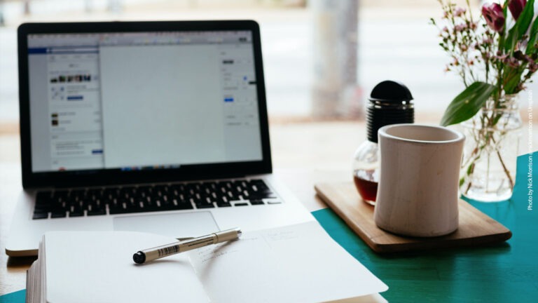 Schreibtisch mit einem Laptop, einem geöffneten Notizbuch mit Stift und einer Teetasse sowie Blumen