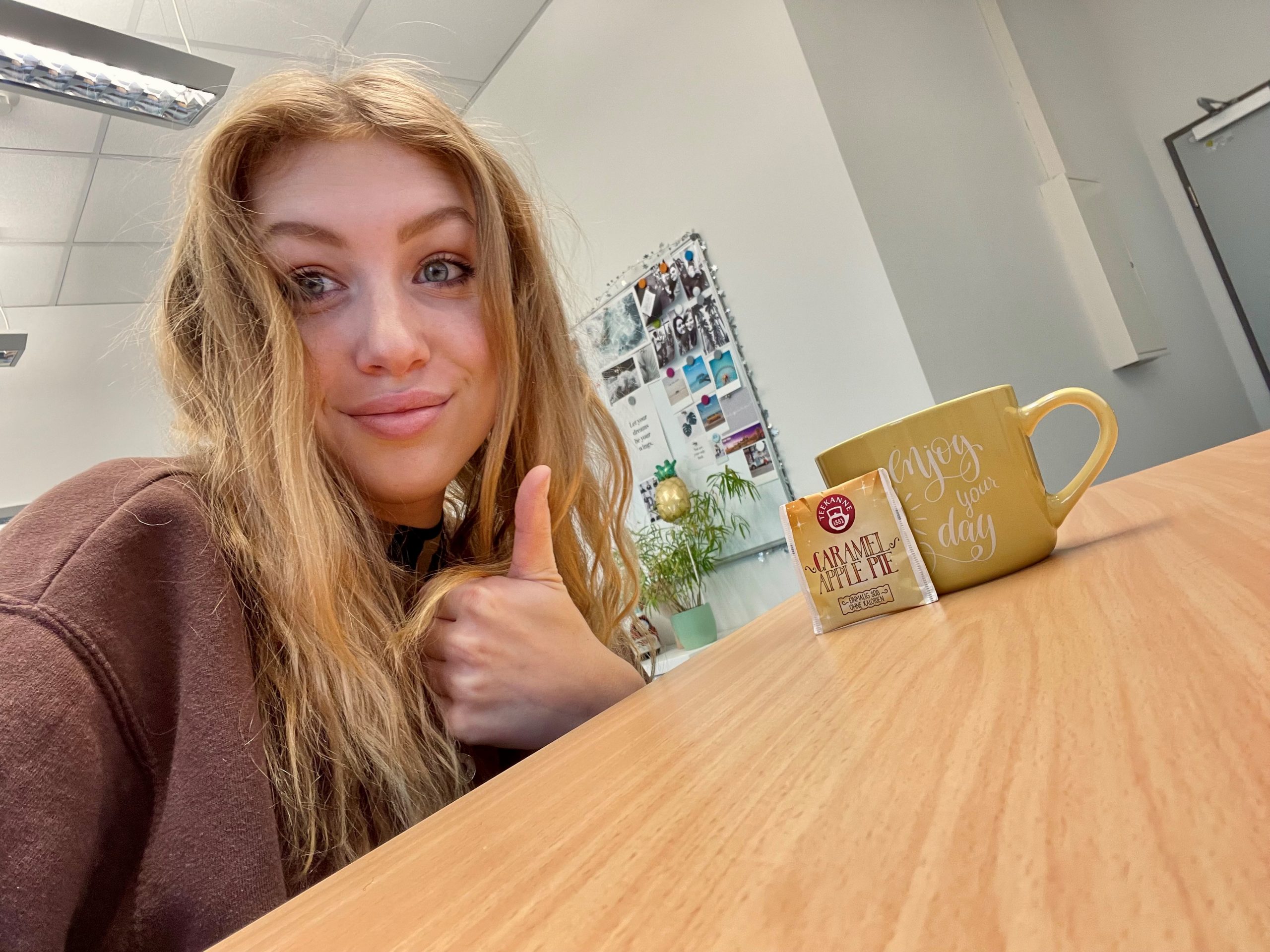 Junge Frau mit blonden Haaren sitzt am Schreibtisch mit einer Tasse Tee, schaut in die Kamera und zeigt einen Daumen nach oben