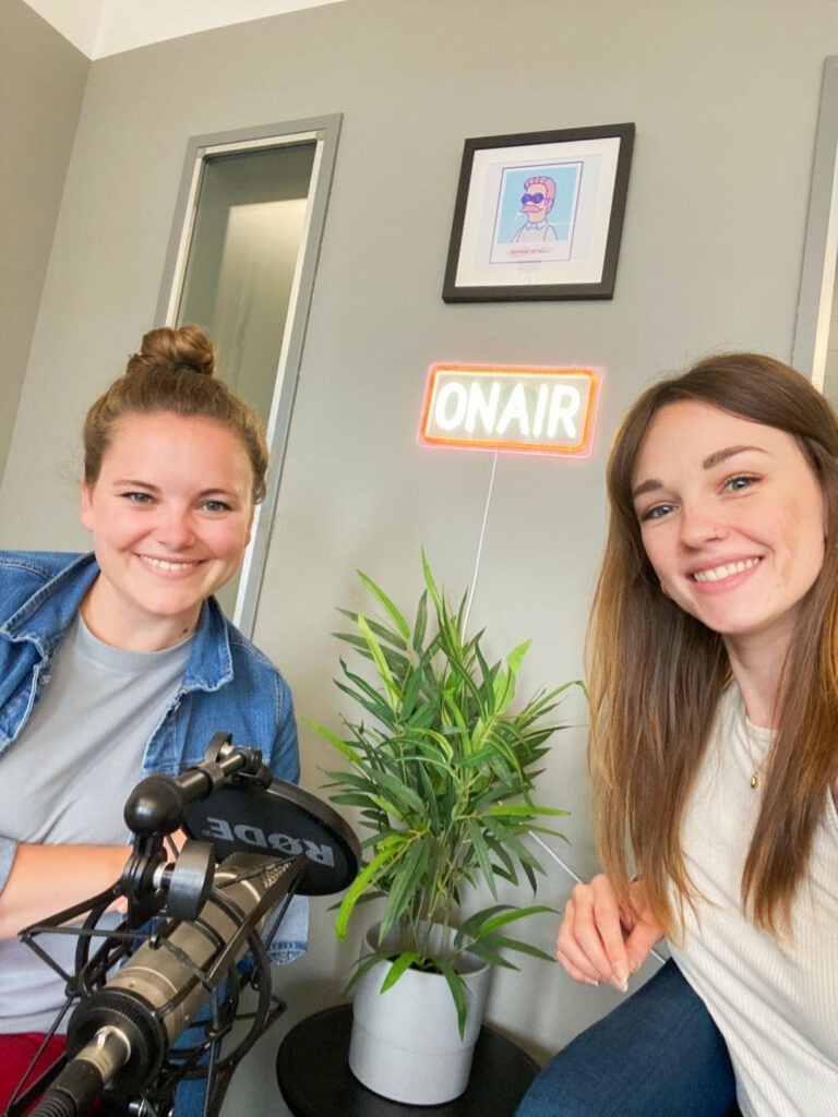 Zwei Frauen sitzen vor einem Podcast-Mikrofon und lächeln in die Kamera