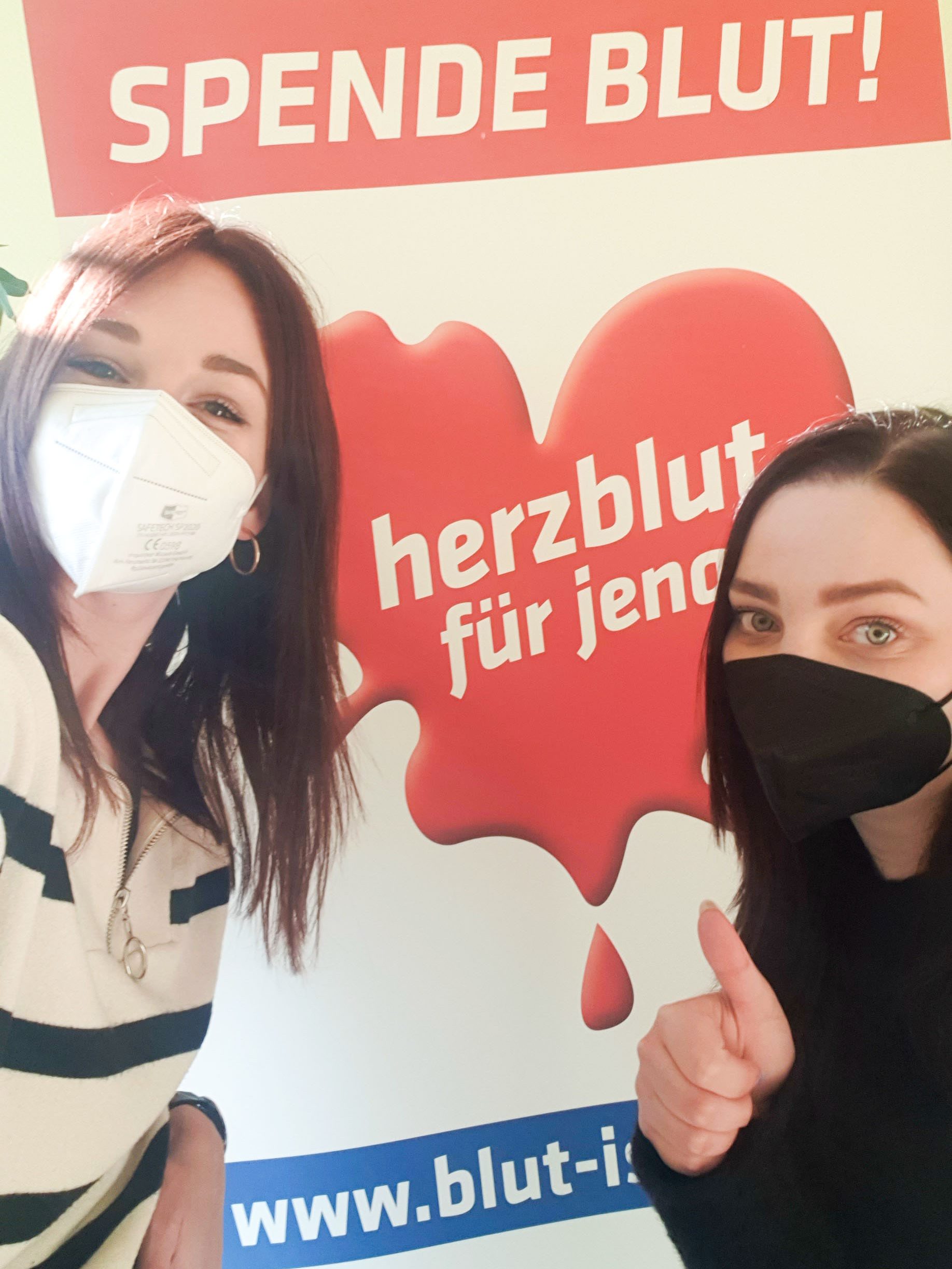 Zwei Frauen mit FFP2-Maske posieren für ein Selfie vor einem Plakat