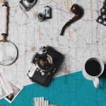 Gegenstände, wie eine Kamera, eine Lupe, eine Kaffeetasse und Polaroidfotos liegen ausgebreitet auf einer Landkarte
