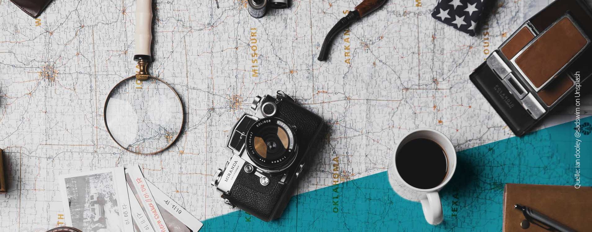 Gegenstände, wie eine Kamera, eine Lupe, eine Kaffeetasse und Polaroidfotos liegen ausgebreitet auf einer Landkarte